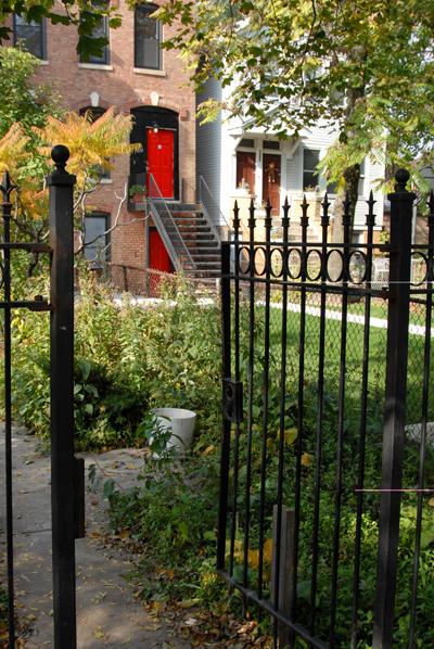 Overgrown front yard of brick home with a red door looking through a black iron gate